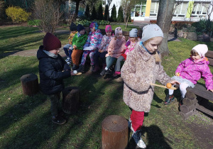 Dwoje dzieci niesie jajka na drewnianych łyżkach. Za nimi na ławkach siedzi siedmioro dzieci.