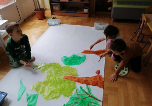 Troje dzieci maluje drzewa na dużym kartonie leżącym na podłodze.