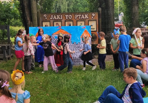 Zdjęcie w ogrodzie przedszkolnym. Dzieci stoją na tle namalowanego zamku. przed nimi leżą maty, na których siedzą inne dzieci.