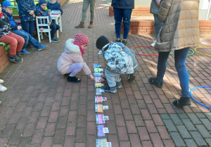 Troje dzieci kuca przy rozłożonych na tarasie obrazkach lokomotywy i wagonów.