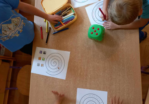 Czworo dzieci przy stoliku wypełnia karty pracy z narysowaną spiralą. Między dziećmi leży kostka do gry i pudełko z kredkami.