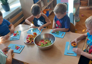 Pięcioro dzieci siedzi przy stoliku. Kroją winogrona i arbuza na deskach do krojenia. Pomiędzy dziećmi stoi talerz z całymi owocami i miska z pokrojonymi owocami.