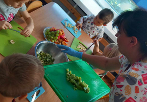 Dzieci kroją owoce na sałatkę. Obok stoi kucharka z deską do krojenia, na której ma pokrojone kiwi.