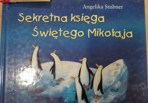 Okładka książki "Sekretna księga Świętego Mikołaja" przedstawiająca pięć pingwinów na krze patrzących na sanie Świętego Mikołaja lecące po nocnym niebie. Na lewym górnym rogu książki leży mała, czerwona książeczka.