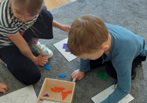 Dzieci na dywanie układają kolorowe fklocki w kształcie figur geometrycznych na kartkach z wzorami.