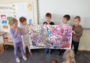 Pięcioro dzieci stoi na dywanie, trzymając wspólnie wykonaną abstrakcyjną pracę plastyczną w dużym formacie.