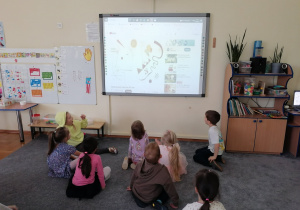Dzieci siedzą na dywanie przed tablicą interaktywną, na której wyświetlana jest animacja o obrazie Wassilego Kandynsky'ego.