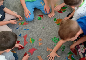 Dzieci siedzą na dywanie i bawią się kolorową mozaiką z trójkątów.