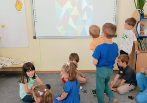 Dzieci siedzą na dywanie. Na tablicy interaktywnej wyświetla się zdjęcie obrazu "Trójkąty i trójkąty" Adama Kały.