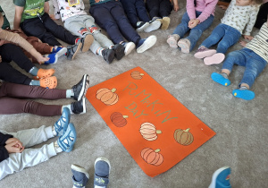 Dzieci siedzą na dywanie dookołapomarańczowego plakatu, na którym jest napis Pumkin Day, otoczony pokolorowanymi i wyciętymi z papieru dyniami.