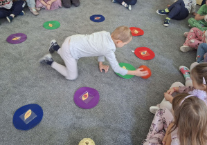 Dzieci siedzą w kole na dywanie. W środku koła chłopiec dotyka jedną z leżących okrągłych plansz.