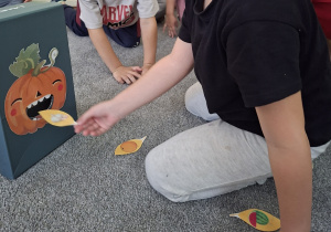 Dziecko wkłada papierowy listek do pudełka z przyklejoną uśmiechniętą dynią.