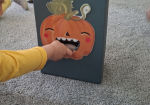 Dziecko trzyma rękę przy otworze w kartonie z przyklejoną uśmiechniętą dynią.