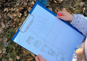 Dziecko trzyma w ręku kartę pracy z symbolami: ławki, pnia drzewa, stołu, kompostownika, tablicy. Przy symbolu ławki zaznaczono 6 pionowych kresek.