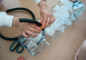 Dzieci umieszczają kawałek gumowego węża w otworze zrobionym w plastikowej butelce i uszczelniają go plasteliną.