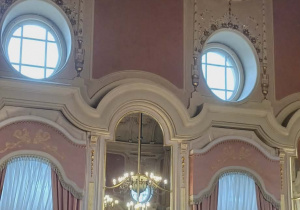 Fragment sali w secesyjnym pałacu z dwoma dużymi oknami i dwoma mniejszymi oknami w kształcie koła. Między oknami stoi zdobiona komoda, a nad nią lustro.