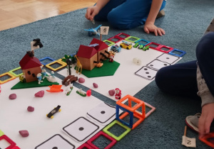Na dywanie leżą dwa białe kartony, otoczone kwadratowymi klockami magnetycznymi. Na każdym kartonie stoi domek i leżą figurki, małe samochodziki i zabawki.