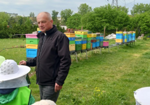 Mężczyzna stoi przodem do dzieci. Za nim widać kolorowe ule.