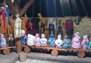 Dzieci siedzą na ławkach, tyłem do fotografa. Przed nimi mężczyzna gra na instrumencie.