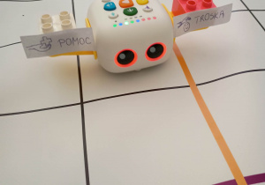 Robot edukacyjny Zosia - mała nauczycielka z przyczepionymi do ramion klockami z napisami troska i pomoc stoi na macie do kodowania.