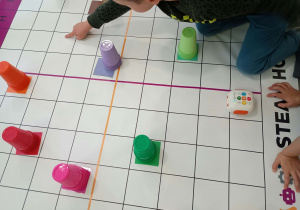 Dziecko wskazuje pole na macie do kodowania, na której stoją kolorowe kubki i robot Zosia.