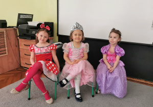 Trzy dziewczynki przebrane za księżniczki siedzą na krzesłach.