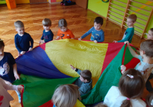 Dzieci trzymają kolorową chustę i zawijają w nią dziecko siedzące w środku.