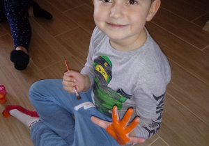 Chłopiec maluje sobie dłoń pomarańczową farbą.