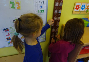 Dwie dziewczynki zawieszają breloczki ze zdjęciami na tablicy wizualizującej współpracę w parach.