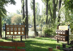 Strefa aktywności w ogrodzie przedszkolnym z dwoma drewnianymi grami, ławkami i pieńkami drzew.